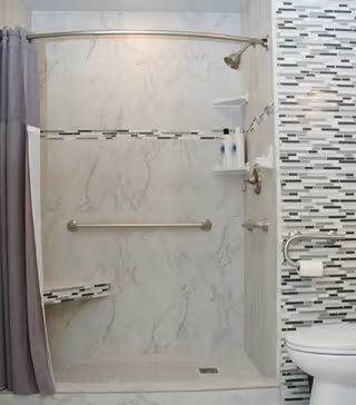 A bathroom shower area with marble-patterned walls, a built-in corner seat, a grab bar, and a curved shower curtain rod with a gray curtain. Adjacent to the shower is a wall with vertical mosaic tiles in shades of gray, white, and black, featuring a toilet with a grab bar and a toilet paper holder.