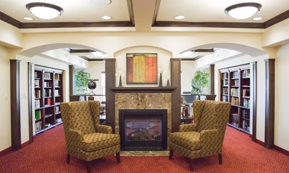 A cozy senior living lounge with two patterned armchairs facing a central fireplace, flanked by bookshelves and decorative arches.