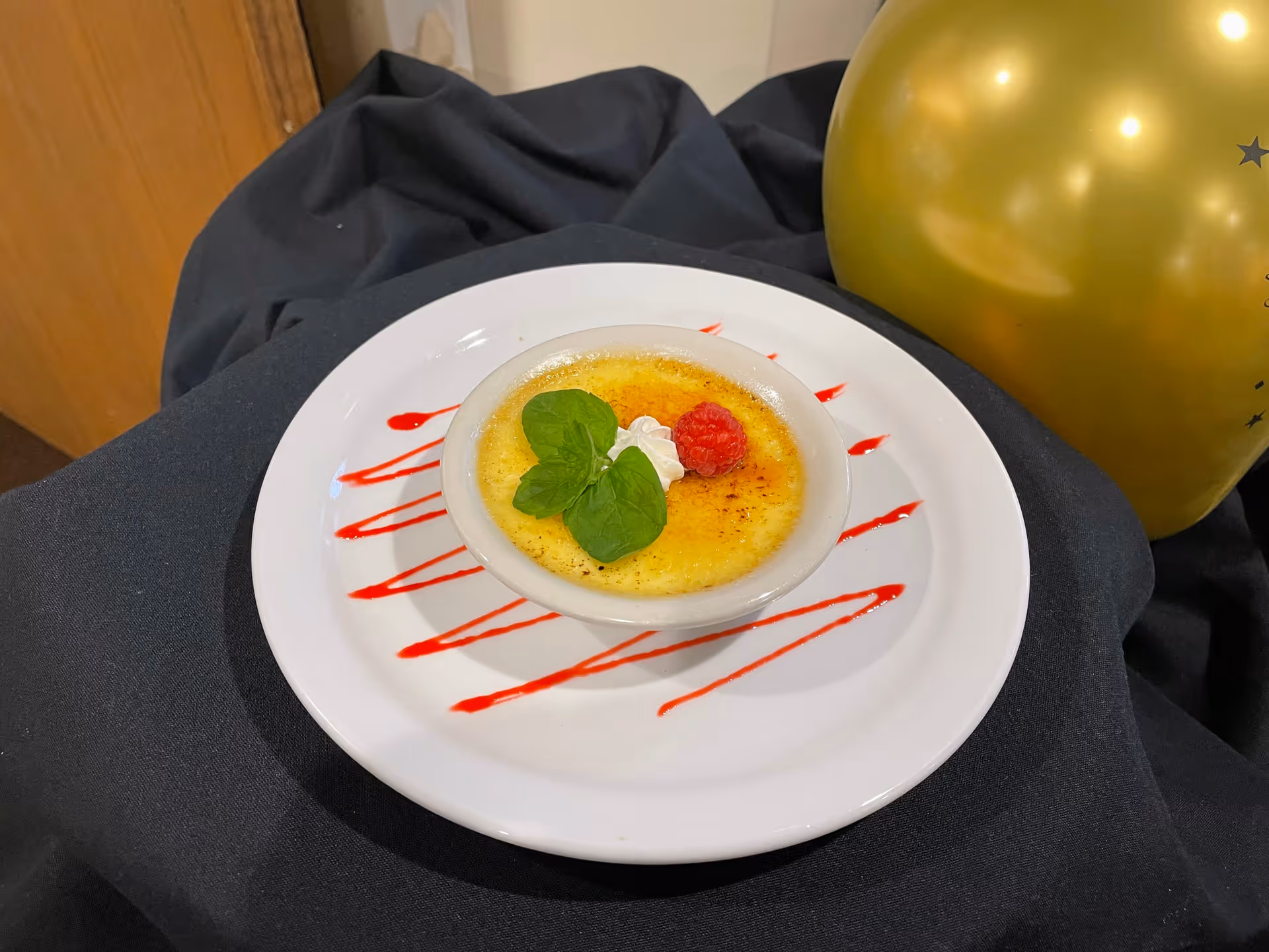 A small dish of crème brûlée garnished with a raspberry, a dollop of whipped cream, and a sprig of mint, served on a white plate with red sauce drizzled in a zigzag pattern. The plate is placed on a black cloth with a gold balloon partially visible in the background.