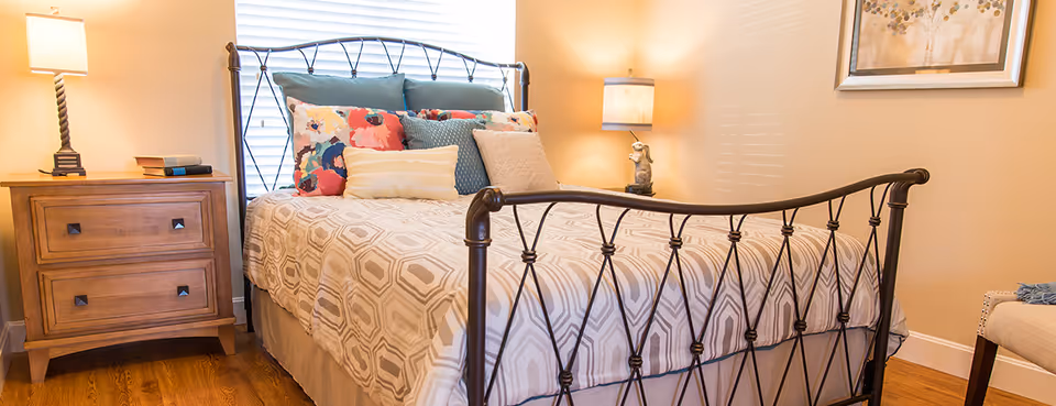 A cozy bedroom with a metal frame bed featuring a patterned beige and white bedspread and multiple colorful pillows. There are two wooden nightstands on either side of the bed, each with a lamp. The room has warm lighting and a framed artwork on the wall.
