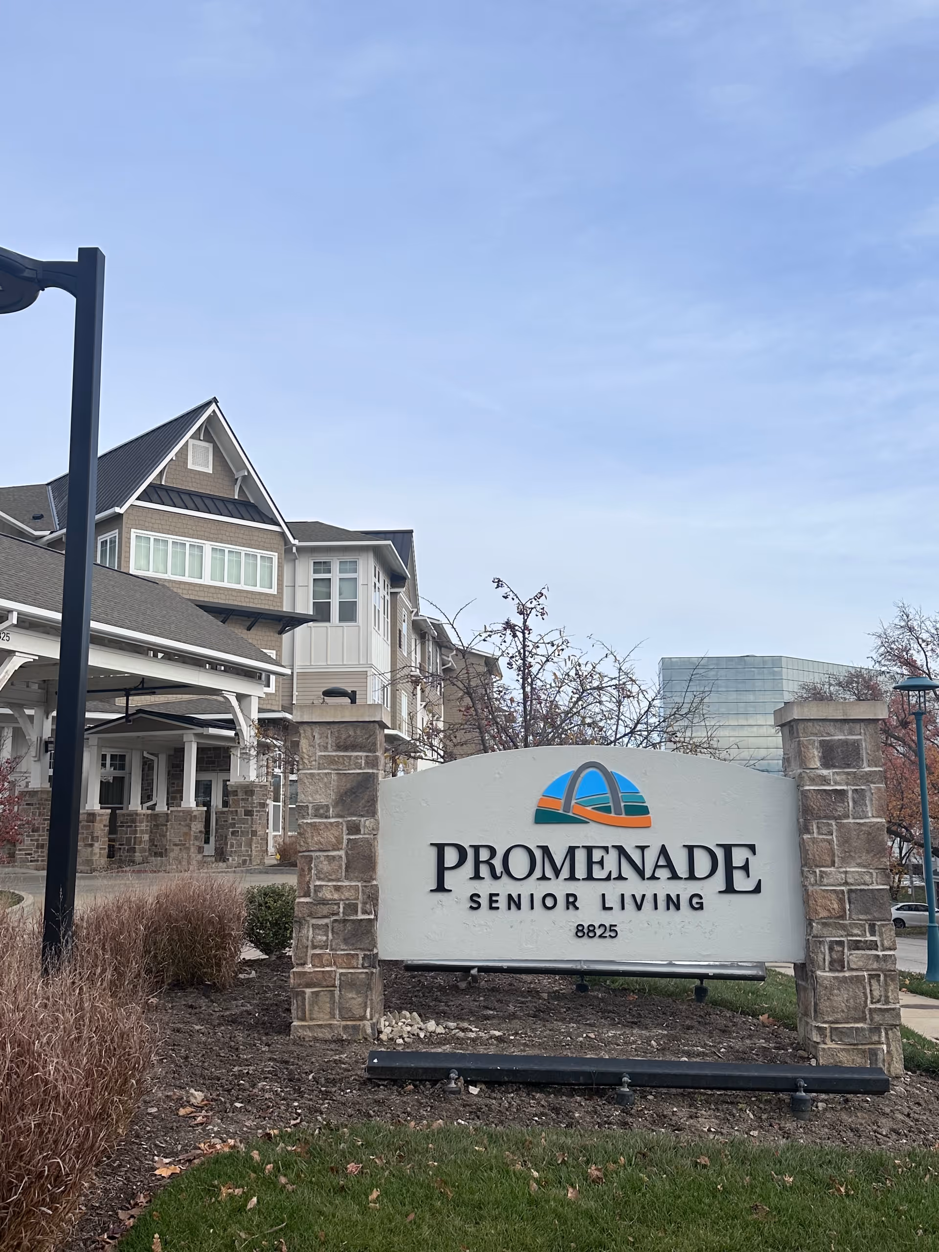 Outdoor view of the Promenade Senior Living facility sign with the building and landscaping in the background under a clear sky.