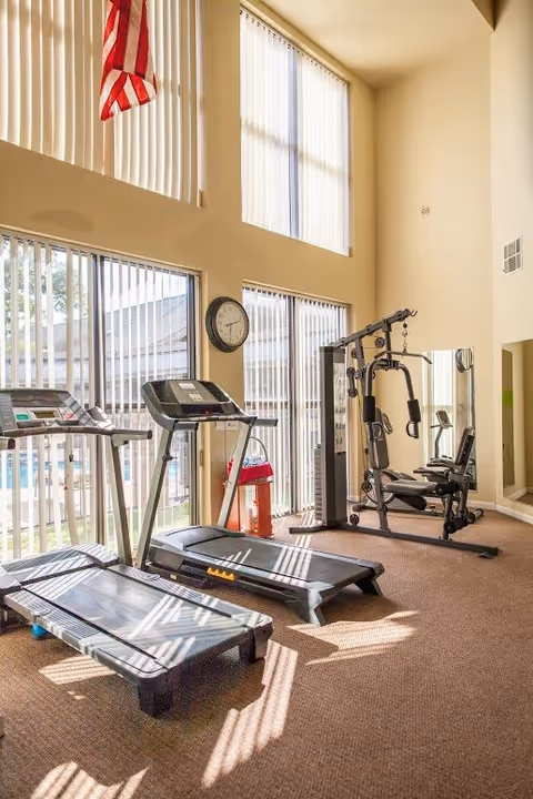 Bright fitness room with two treadmills, a multi-station weight machine, large windows with vertical blinds and an American flag overhead.