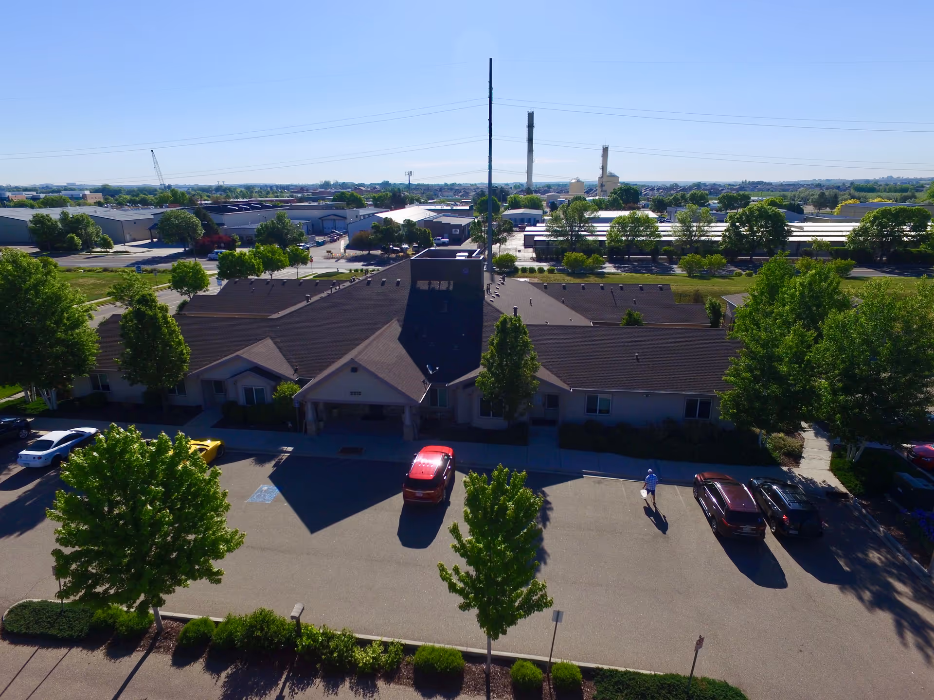 Aerial view of a single-story building with a dark roof surrounded by trees and parked cars in front. The building is part of a larger complex with industrial buildings and greenery in the background under a clear blue sky.