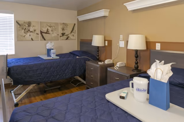 Shared resident room with two hospital-style beds with navy quilts, bedside tables, lamps and overbed trays.