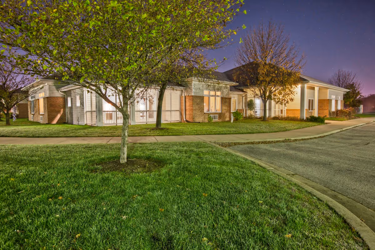Exterior view of a senior living facility building at night with well-maintained green grass, trees with some leaves, a sidewalk, and a paved driveway under a clear sky.