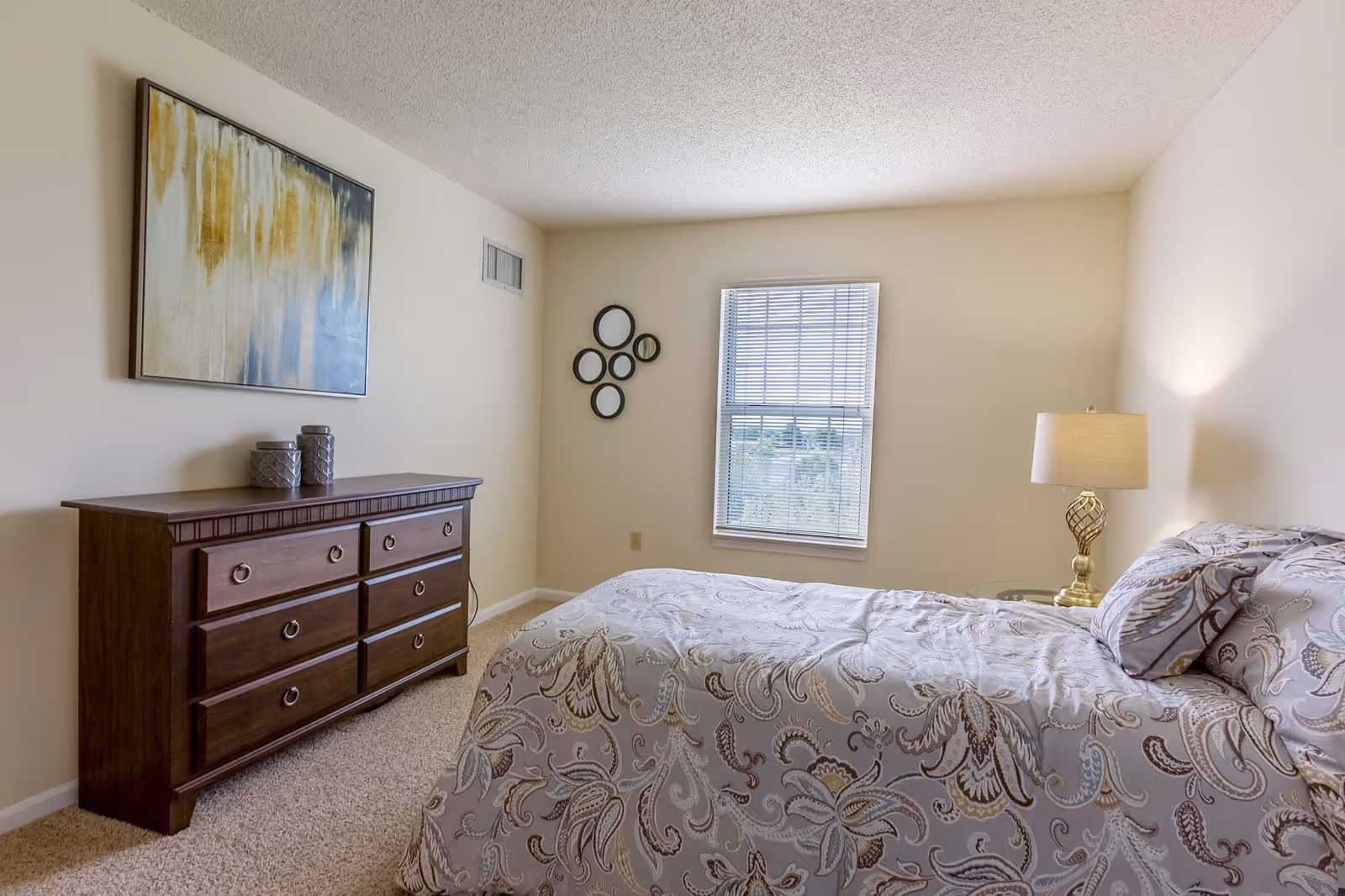 A bedroom with a bed covered in a patterned comforter and matching pillows. There is a wooden dresser with six drawers on the left side, topped with two decorative jars. Above the dresser hangs an abstract painting with yellow, white, and blue tones. A window with blinds is centered on the far wall, and a small round mirror arrangement is mounted to the left of the window. A bedside table with a decorative lamp is on the right side of the bed. The room has beige walls and carpeted flooring.