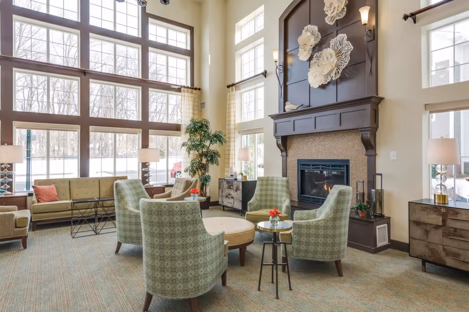 A bright and spacious living room area with large floor-to-ceiling windows letting in natural light. The room features a fireplace with decorative wall art above it, surrounded by four patterned armchairs and a round ottoman in the center. There are two side tables with small red flower arrangements, lamps on wooden dressers, and a green sofa with a coral pillow near the windows. A tall indoor plant is placed near the windows, and the room has a cozy and inviting atmosphere.