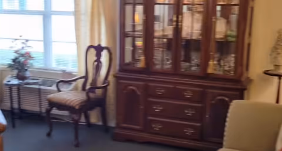 A cozy interior room featuring a wooden china cabinet, an upholstered armchair by a window, and a small side table with flowers.
