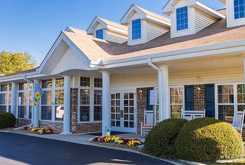 Exterior view of a senior living facility building with brick and beige siding, multiple large windows, white columns, and a covered entrance. There are colorful flower beds and neatly trimmed bushes in front of the building under a clear blue sky.