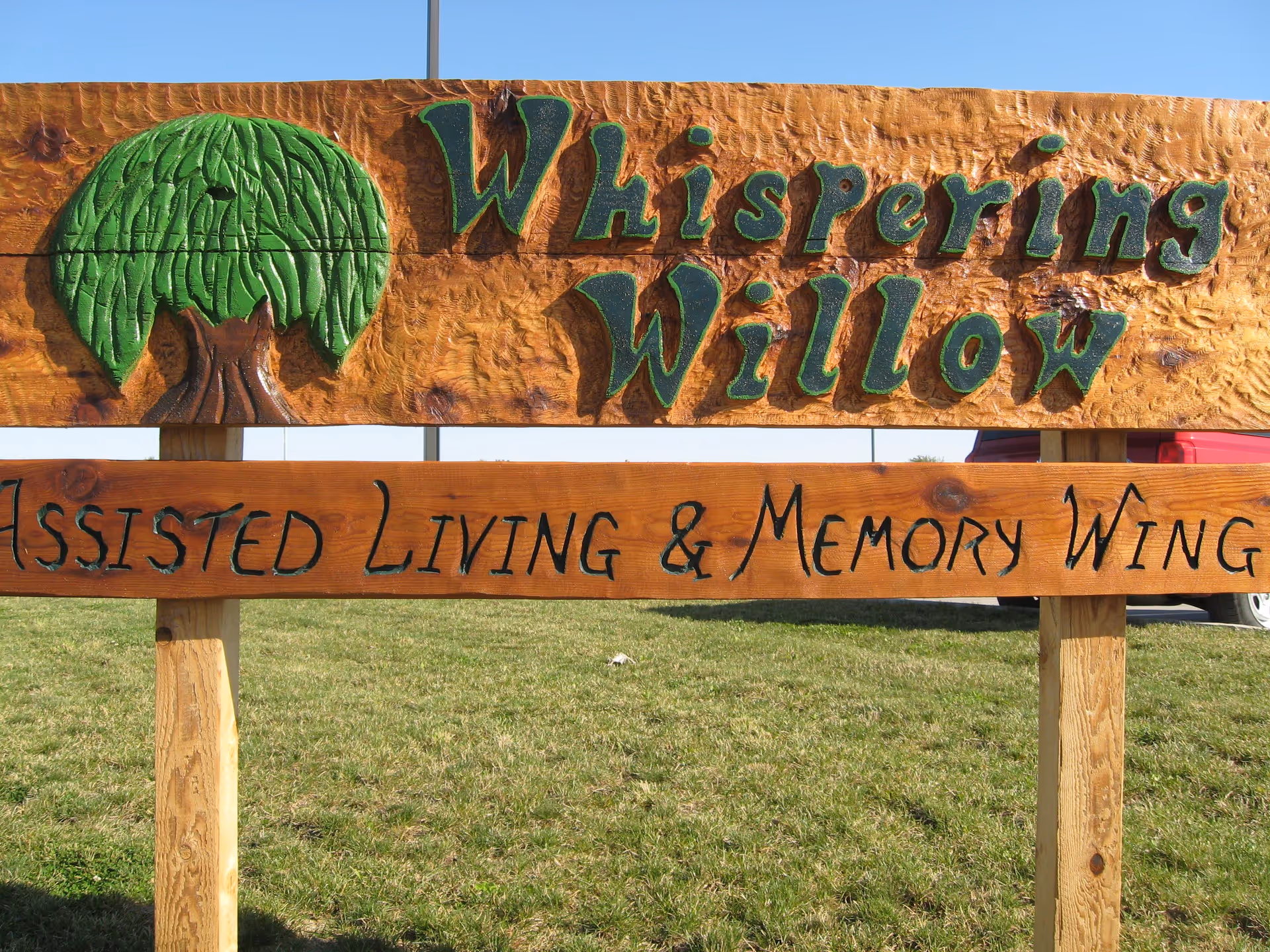 Carved wooden sign reading 'Whispering Willow Assisted Living & Memory Wing' with a painted green willow tree on a grassy lawn.