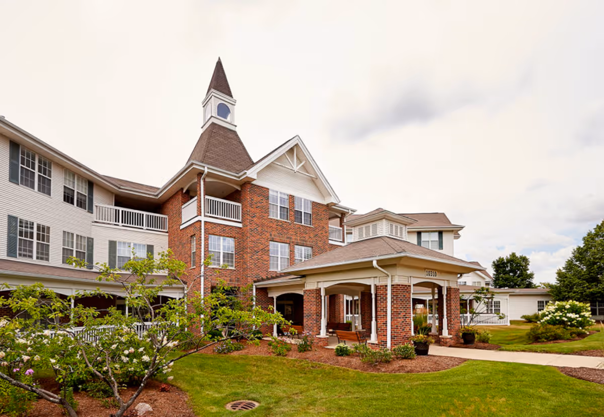 Exterior view of a senior living facility named Peace Village featuring a multi-story building with a brick and white siding facade, a peaked roof with a small tower, landscaped garden with green grass, bushes, and trees, and a covered entrance with benches.