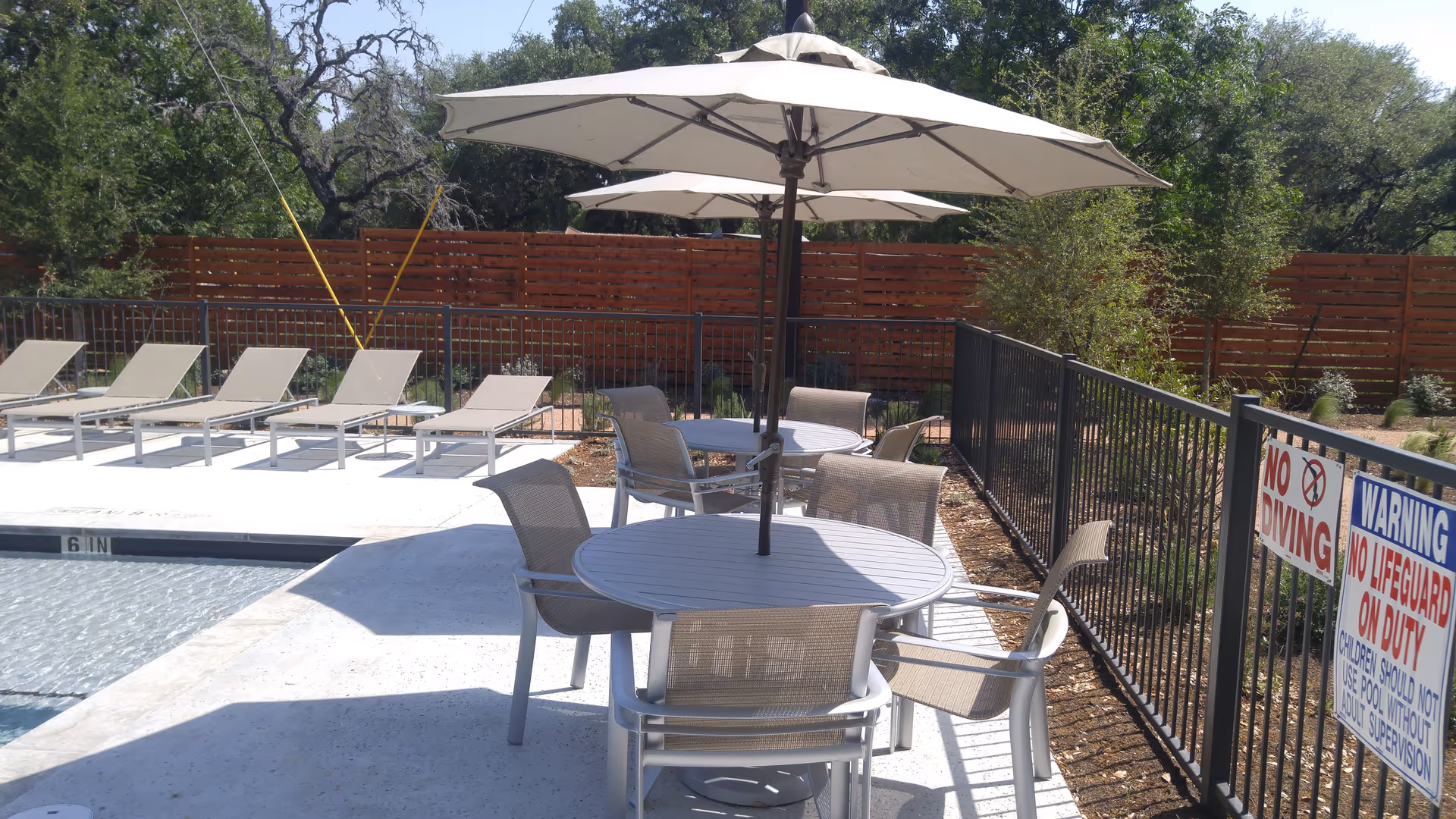 Outdoor pool area with several lounge chairs lined up on the left side and two round tables with umbrellas and chairs on the right side. The area is enclosed by a black metal fence with warning signs about no diving and no lifeguard on duty. There is a wooden privacy fence and trees in the background.