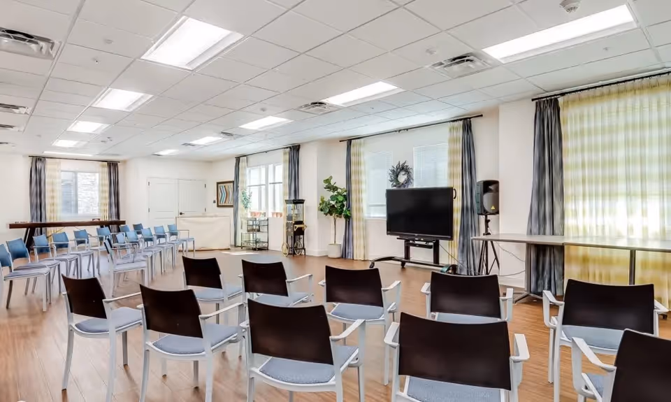 Bright multipurpose activity room with rows of chairs facing a television, large windows with curtains, and wood flooring.