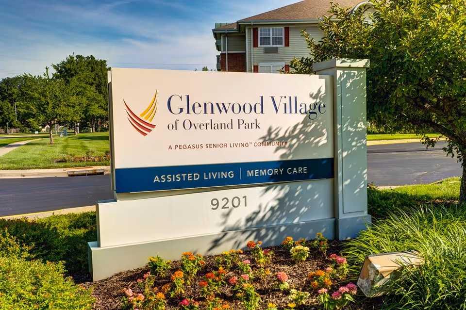 Outdoor sign for Glenwood Village of Overland Park, a Pegasus Senior Living community offering assisted living and memory care, surrounded by greenery and flowers with a building and road in the background.