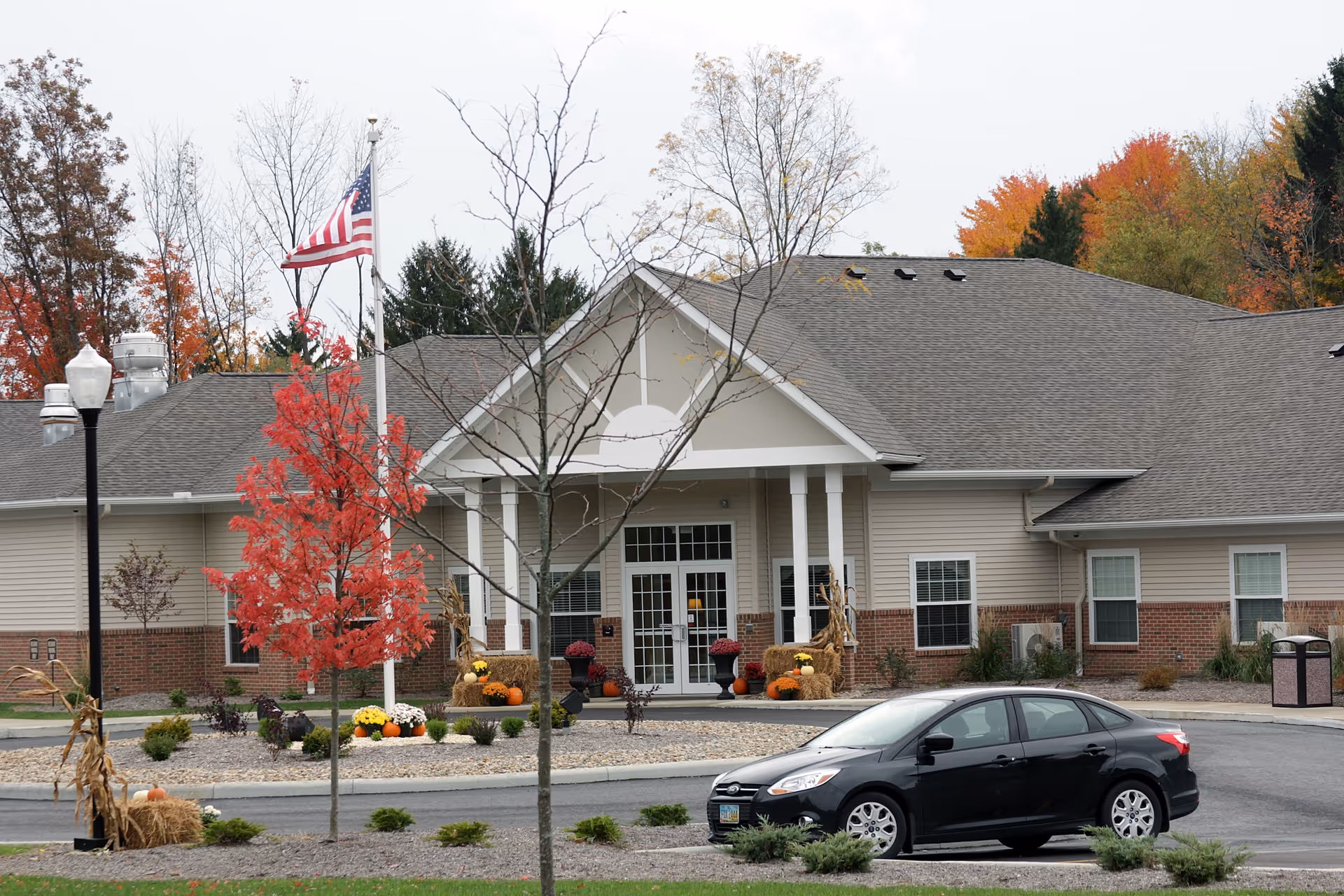 Exterior view of Danbury Wooster facility showing the entrance with a covered porch supported by white columns. There is a flagpole with an American flag, autumn decorations including pumpkins and hay bales, and a black car parked in front. Trees with fall foliage are visible in the background.