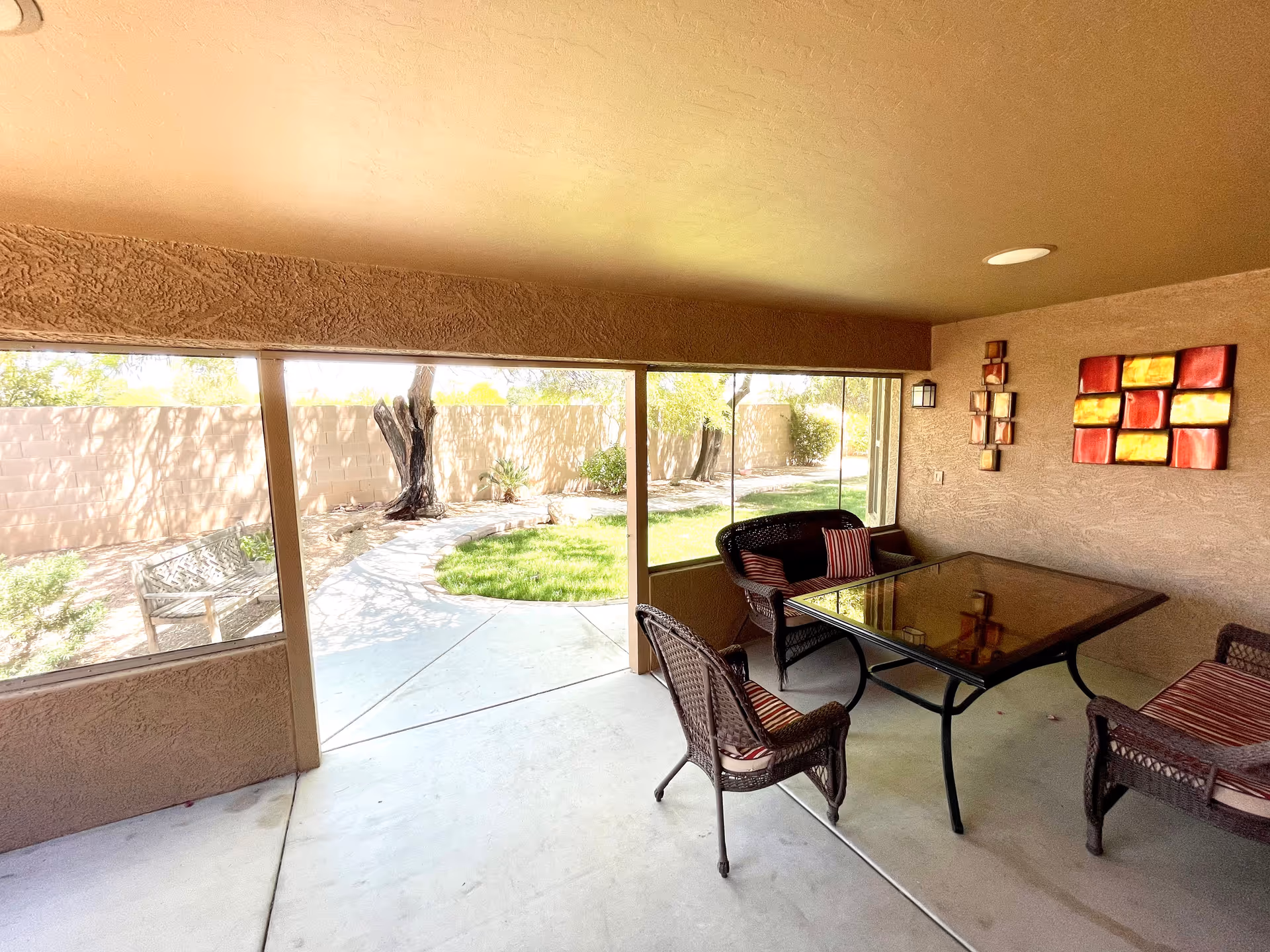 Covered patio area with a glass-top table surrounded by four wicker chairs with cushions. The patio has textured beige walls decorated with colorful square wall art. Large windows provide a view of a garden with a curved concrete path, green grass, trees, and a wooden bench outside.