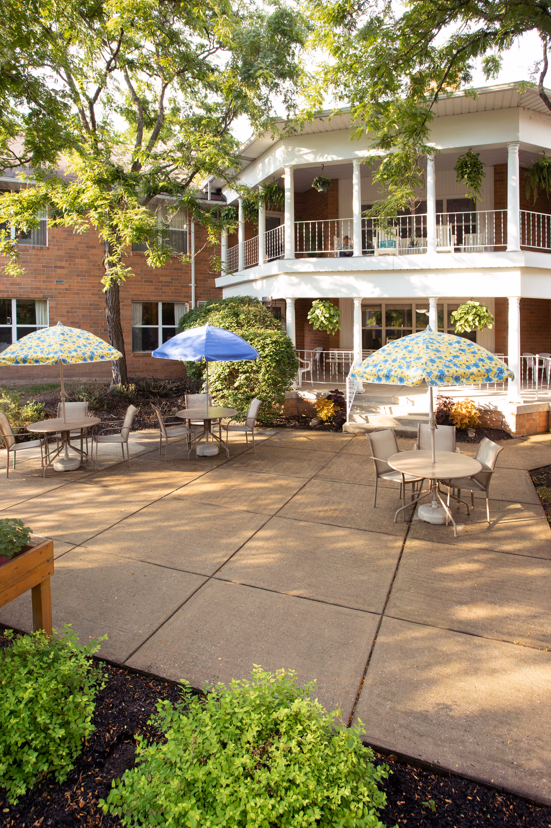 Outdoor patio area at Royalton Woods with several round tables and chairs, each shaded by colorful umbrellas. The patio is surrounded by greenery and trees, with a two-story brick building featuring white railings and hanging plants in the background.