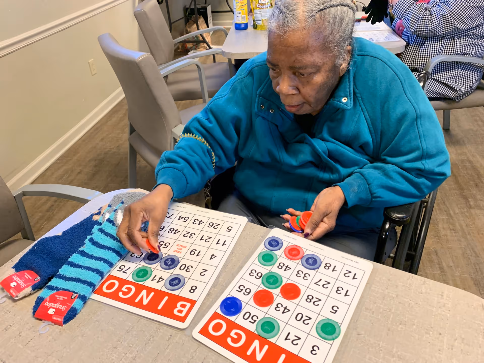 An elderly woman in a wheelchair playing bingo at a table with two bingo cards and colorful markers. A pair of blue striped socks is also on the table.