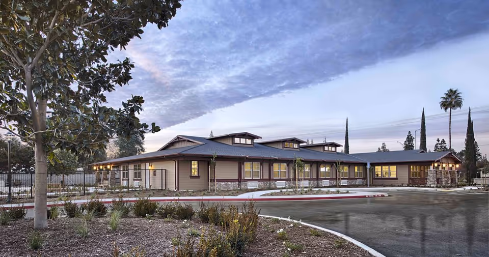 Exterior view of Magnolia Crossing Assisted Living facility during dusk, showing a single-story building with multiple windows, a landscaped area with trees and shrubs, and a curved driveway in front.