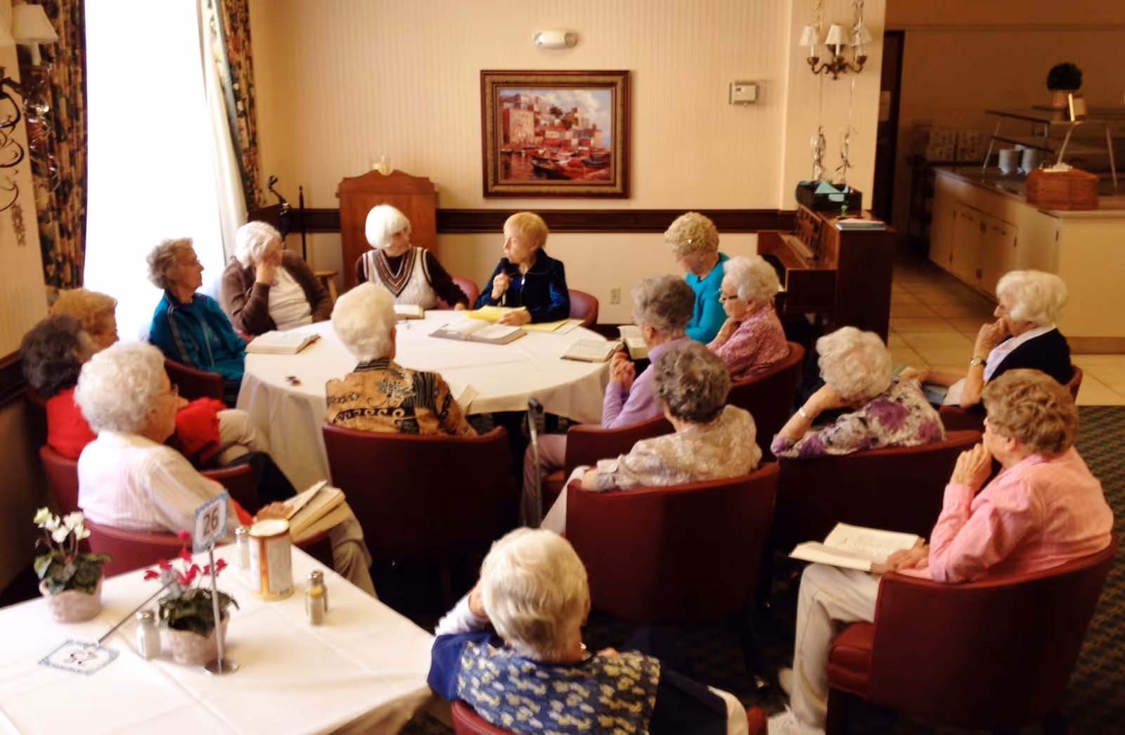 A group of elderly women seated in a circle around a table in a well-lit room, engaged in a discussion or meeting. The room has a painting on the wall, a piano, and a kitchen area visible in the background.