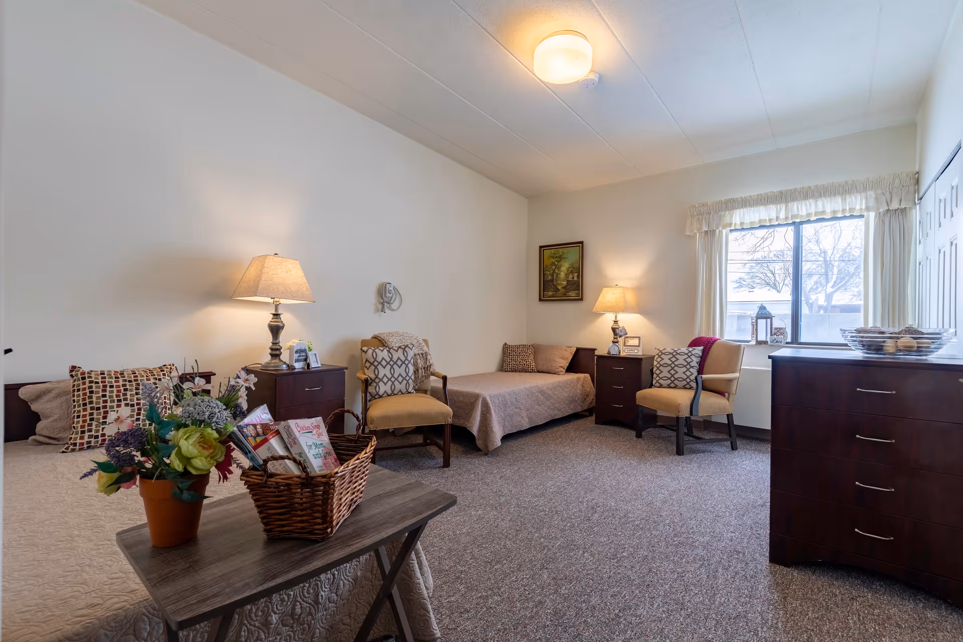 A cozy senior living bedroom with two single beds, two armchairs, two bedside tables with lamps, a wooden dresser, a window with curtains, and a small table with a flower pot and a basket of books and magazines.