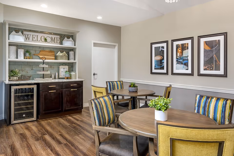 A cozy dining area with two round wooden tables, each surrounded by four chairs with striped and yellow upholstery. On the tables are small potted plants. Against the wall is a dark wood cabinet with a wine cooler on the left side and open shelves above it displaying decorative items, including a 'WELCOME' sign. The walls are painted in neutral tones with three framed pictures hanging on one wall. The floor is wood with a warm finish.