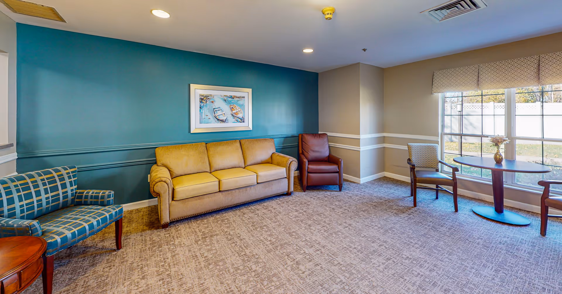 A cozy sitting area in a senior living facility with a mustard yellow sofa, a blue patterned armchair, a brown leather armchair, and a small round table with two chairs near a large window. The walls are painted teal and beige with white trim, and a framed picture of boats hangs above the sofa.