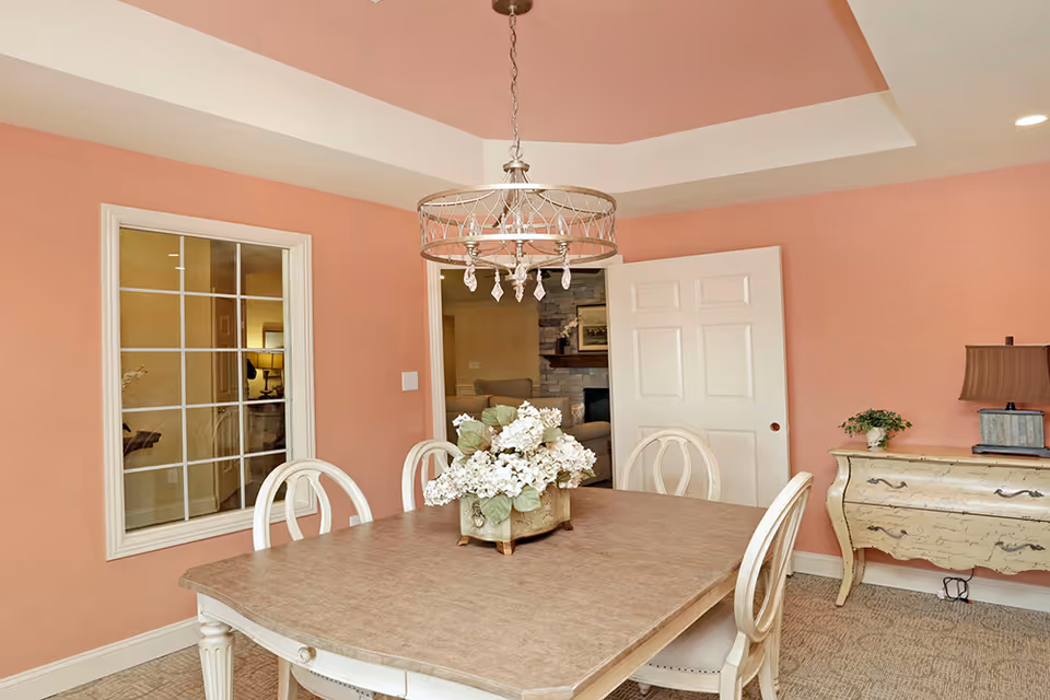 Dining room with a rectangular table, white chairs, a floral centerpiece, chandelier, and pink walls with a sideboard and open doorway.