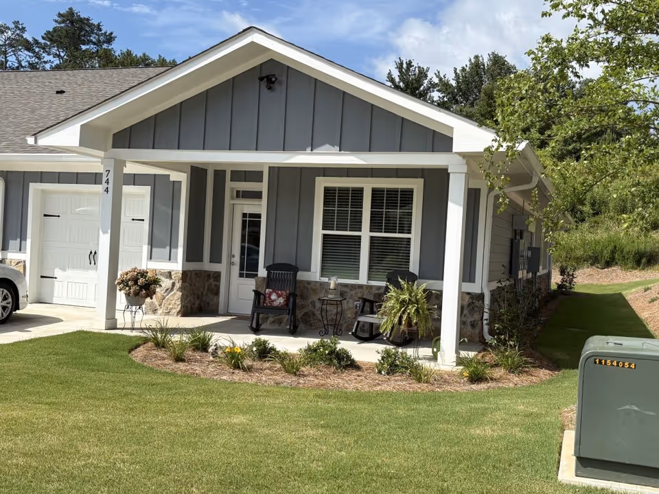 Exterior view of a single-story residential building with gray siding and white trim. The front porch has two black rocking chairs with cushions, a small table with a candle, and potted plants. The lawn is well-maintained with green grass and landscaped flower beds. A garage door is visible on the left side, and a green utility box is on the right side near the sidewalk.