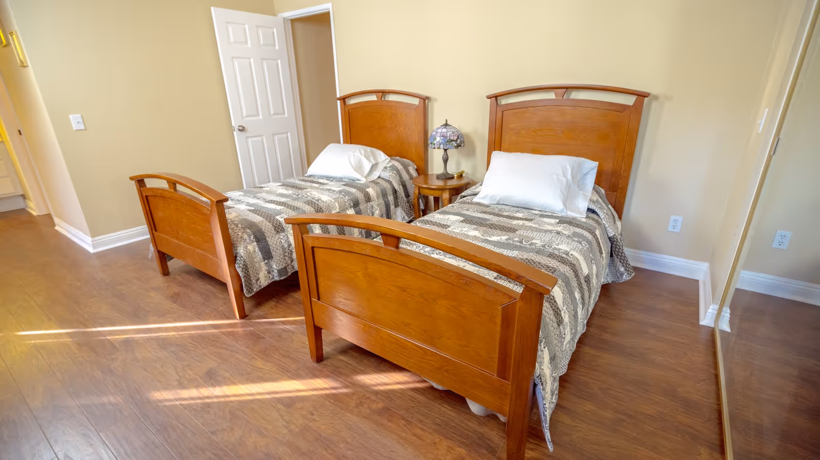 A bedroom with two single wooden beds placed side by side, each with a white pillow and a patterned gray and white quilt. Between the beds is a small round wooden nightstand with a decorative lamp. The room has light beige walls, wooden flooring, a white door, and a mirrored closet door on the right side.
