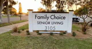 Outdoor image of a white sign with black text that reads 'Family Choice Senior Living 3105' placed on a grassy area near a sidewalk and street, with trees and a parked vehicle in the background during sunset.