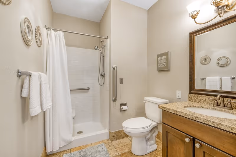 A clean and well-lit bathroom featuring a walk-in shower with a white curtain, a white toilet, a granite countertop with a sink, a large framed mirror, and beige tiled floor. The walls are painted light beige and decorated with two round wall hangings and a small sign that reads 'Grateful, Thankful, Blessed'. There are white towels hanging on a towel rack.