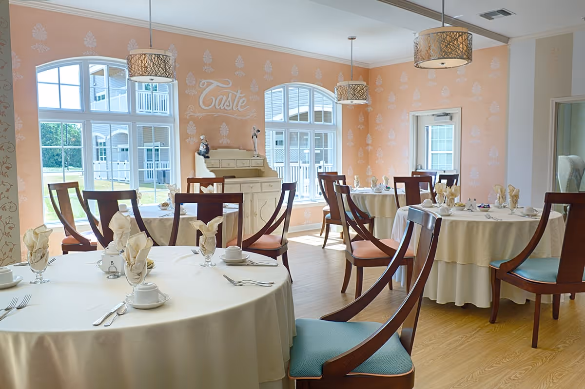 A bright dining room with round tables covered in white tablecloths, each set with folded napkins, cups, and silverware. The room has peach-colored walls with a decorative pattern and large windows letting in natural light. There are wooden chairs with blue and orange cushions around the tables, and three pendant lights hanging from the ceiling. The word 'Taste' is written on one wall above a white sideboard.