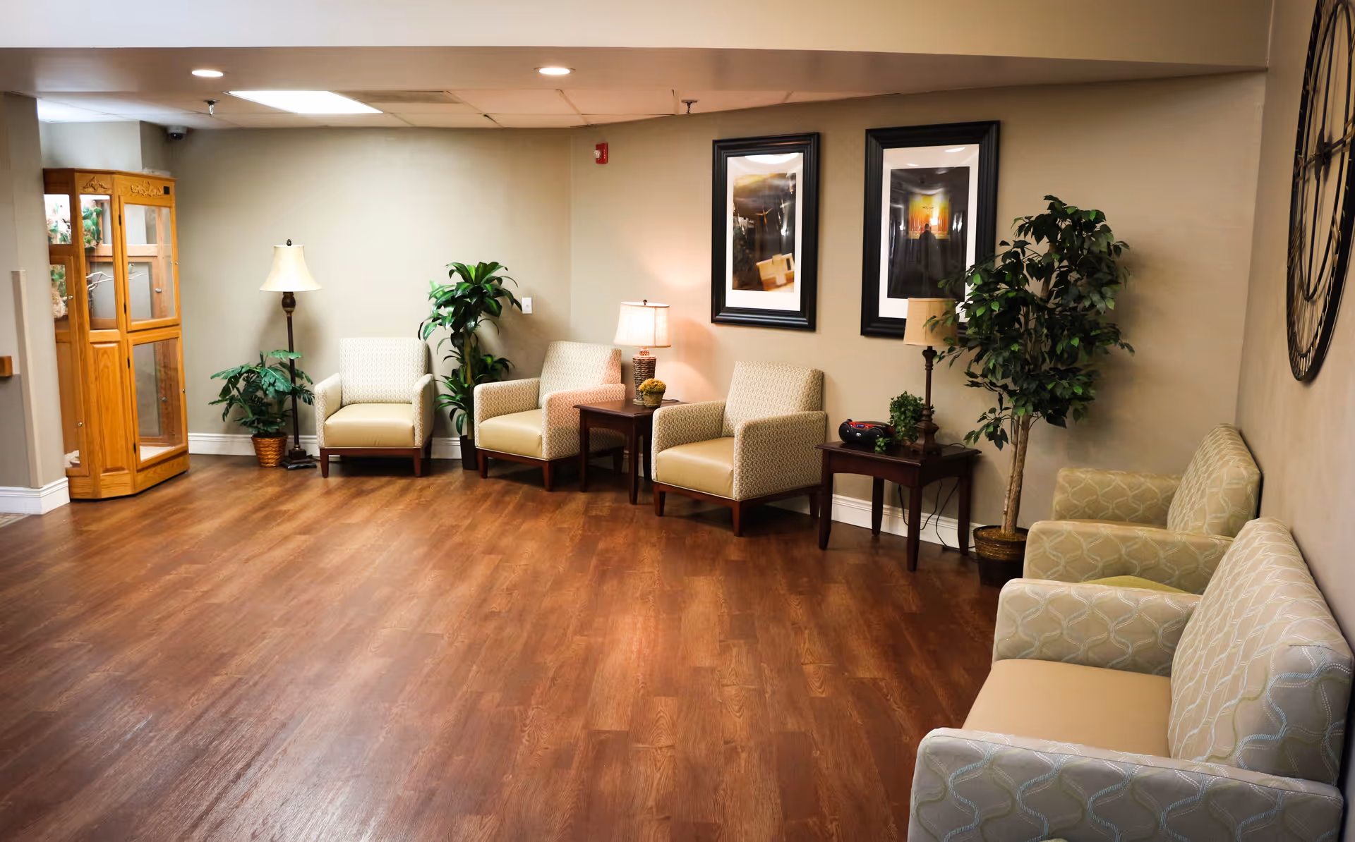 A cozy waiting or sitting area in a senior living facility with several cushioned armchairs arranged along the walls, wooden side tables with lamps, potted plants, framed artwork on the wall, a wooden display cabinet, and wood flooring.