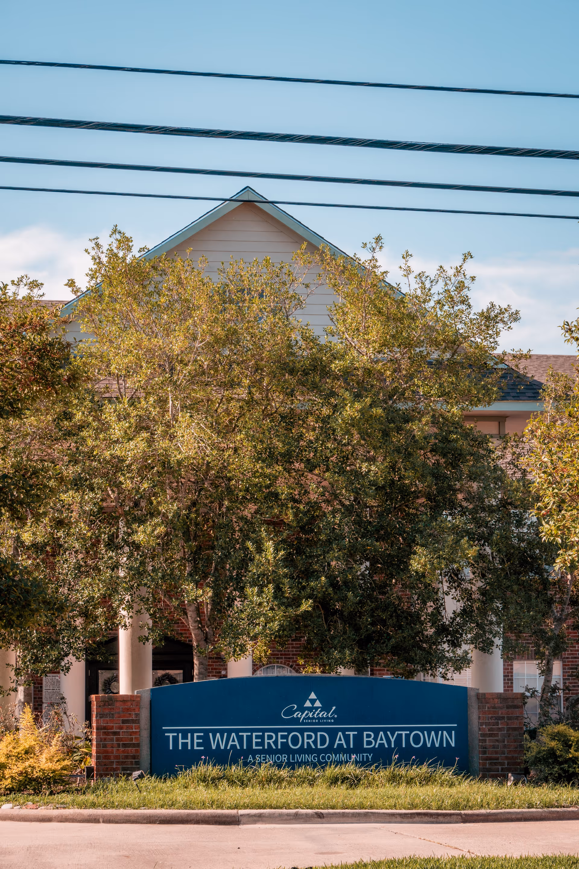 Exterior view of The Waterford at Baytown senior living community sign with trees and part of the building visible in the background under a clear blue sky.