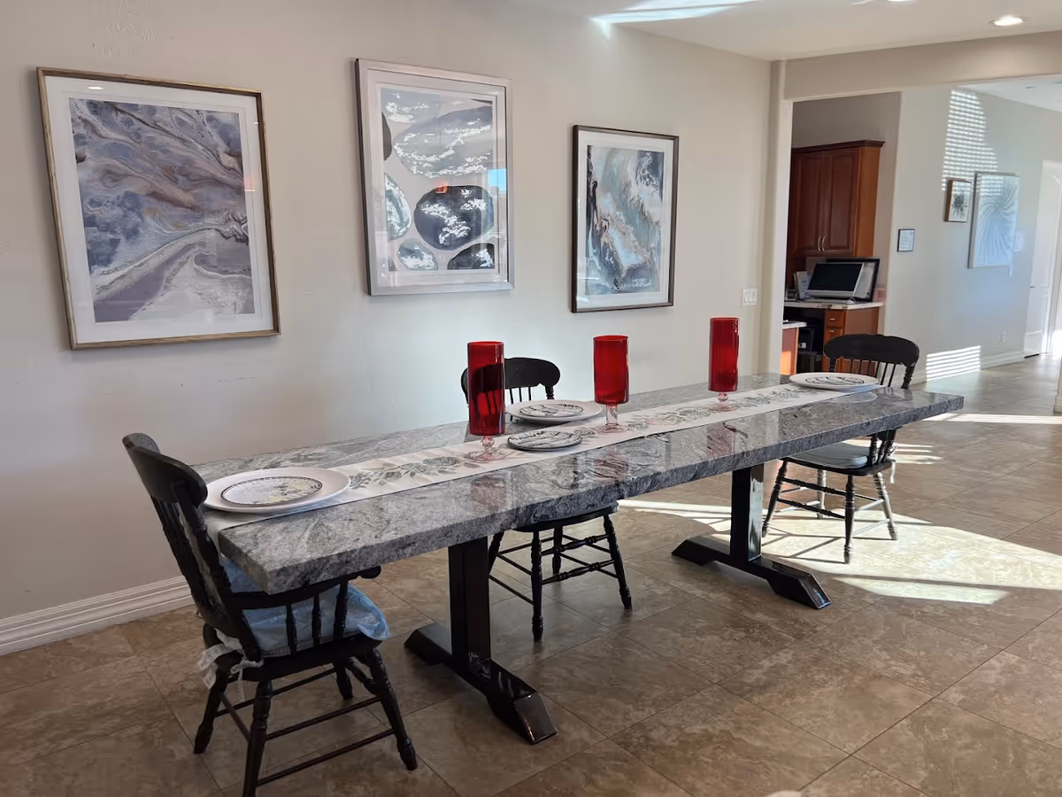 A dining area with a long rectangular marble table set with three red glasses and three plates. There are three black wooden chairs around the table. The wall behind the table has three framed abstract art pieces. The floor is tiled, and there is a hallway visible in the background with additional artwork on the walls.