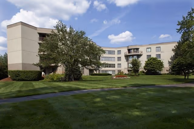 Exterior view of a multi-story retirement community building with beige walls, surrounded by green grass, trees, and a clear blue sky with some clouds.