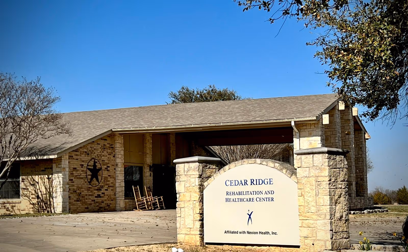 Exterior view of Cedar Ridge Rehabilitation and Healthcare Center building with a stone sign in front displaying the facility name and affiliation with Nexion Health, Inc. The building has a stone and brick facade with a covered entrance and rocking chairs visible on the porch.