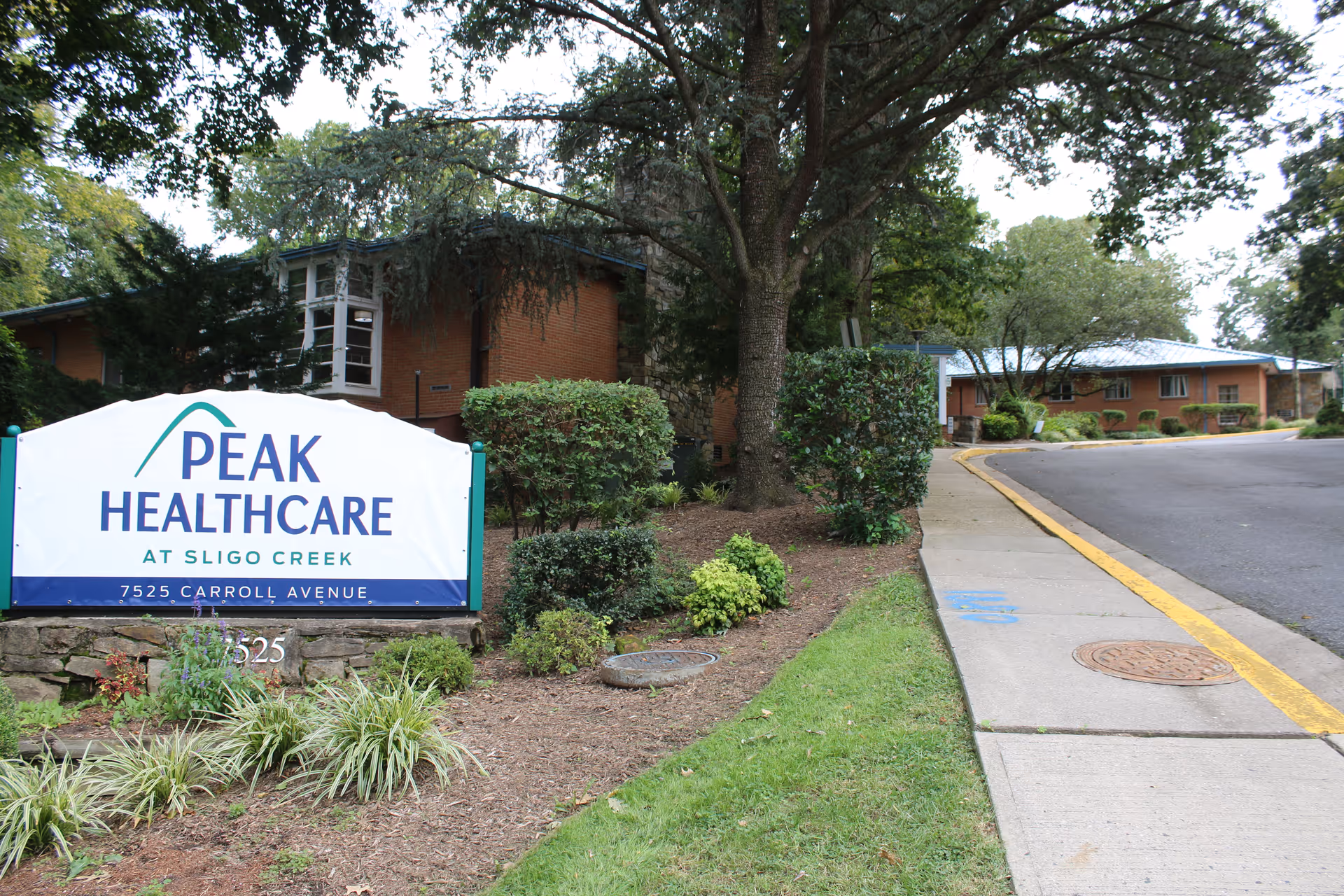 Exterior view of Peak Healthcare at Sligo Creek facility showing a large white sign with the facility name and address, surrounded by landscaped greenery, trees, and a sidewalk leading to the building.