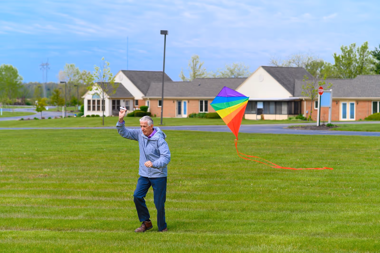 An elderly man flying a colorful rainbow kite on a large grassy field with a senior living community building in the background under a partly cloudy sky.