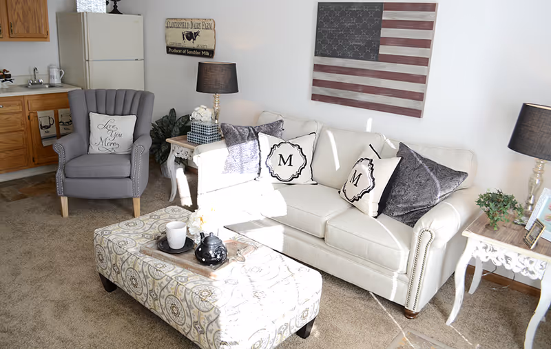 Sunlit living room with a beige sofa, patterned ottoman coffee table, gray armchair, and a small kitchenette in the background.