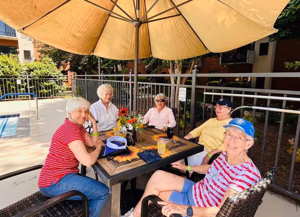 Five elderly women sitting around a square outdoor table with a sunflower design under a large beige patio umbrella. They are smiling and enjoying a sunny day near a swimming pool, with a metal fence and greenery in the background.
