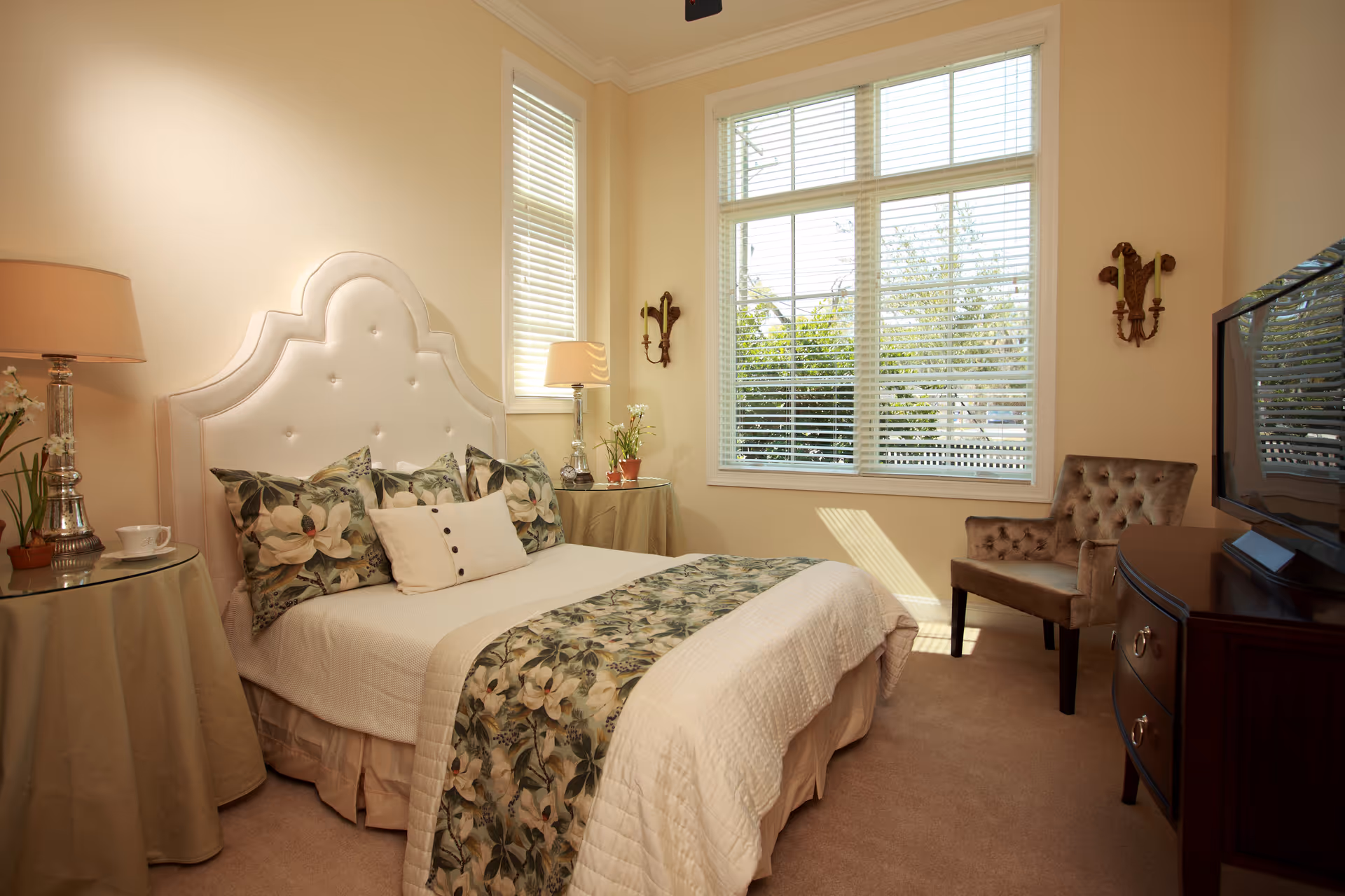 Sunlit bedroom with a bed featuring floral bedding and an upholstered headboard, nightstands with lamps, a tufted chair, and a TV on a dresser.