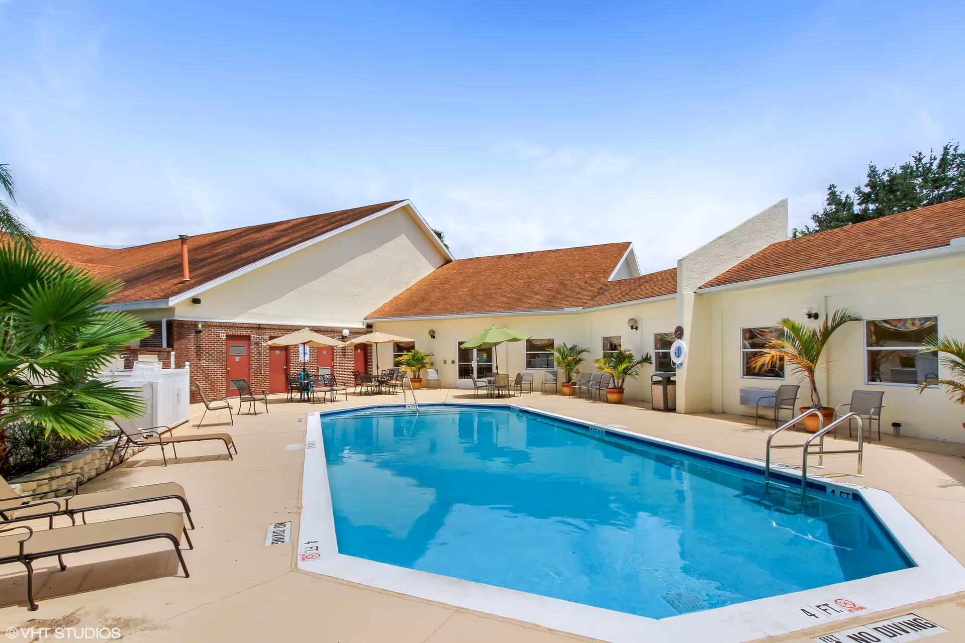 Outdoor swimming pool area at Merrill Gardens at Barkley Place with lounge chairs, tables with umbrellas, potted plants, and a building with red doors and windows surrounding the pool under a partly cloudy blue sky.