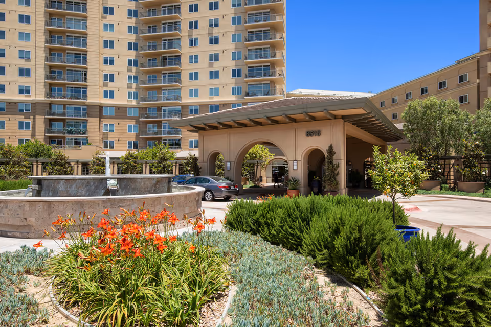 Exterior view of Vi at La Jolla Village showing the entrance with a covered driveway, a circular water fountain, landscaped garden with orange flowers and green shrubs, and a multi-story residential building in the background under a clear blue sky.