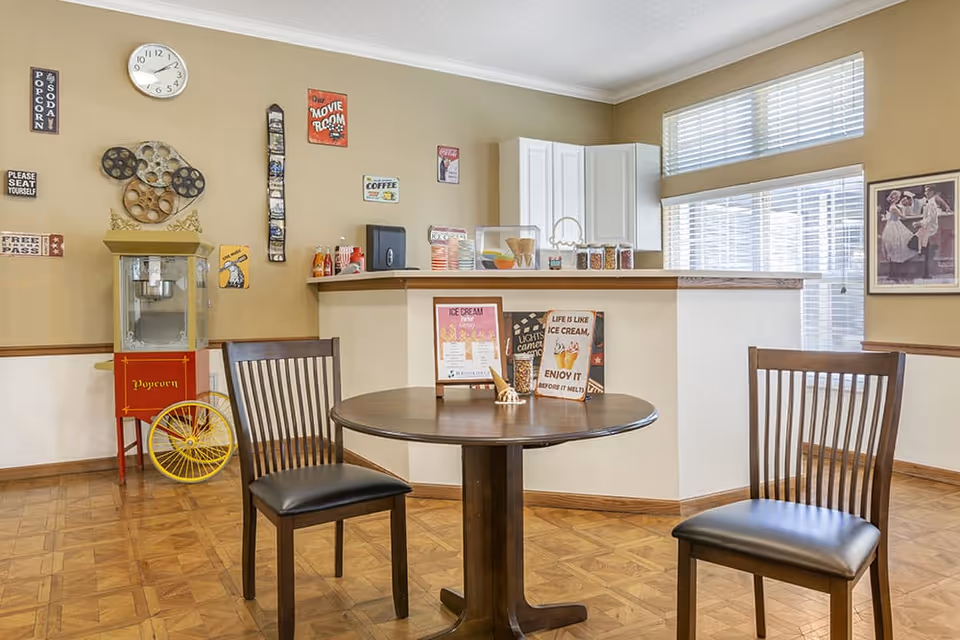 A cozy room with a round wooden table and two wooden chairs with black cushions. Behind the table is a counter with various items including ice cream cones and jars. On the left side, there is a vintage-style popcorn machine with a yellow wheel. The walls are decorated with movie-themed signs, a clock, and a framed picture. Large windows with blinds allow natural light into the room.