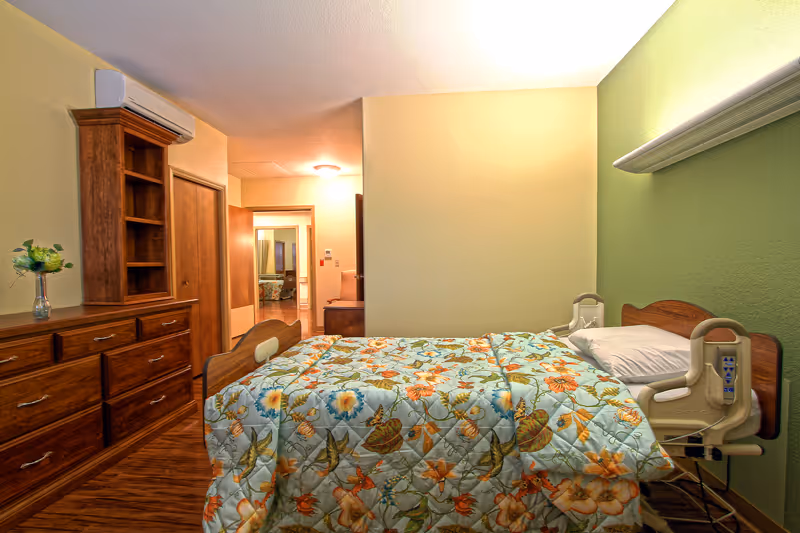 Private bedroom in a care facility featuring an adjustable bed with a floral quilt, wooden dresser and shelving, and a view into a hallway.