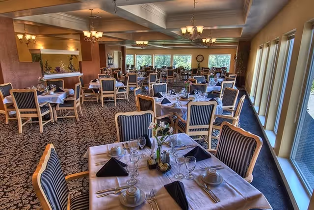 A spacious dining room with multiple tables set for a meal. Each table is covered with a white tablecloth and has neatly arranged place settings including plates, cups, glasses, silverware, and black folded napkins. The chairs have wooden frames with striped upholstery. Large windows line one side of the room, allowing natural light to fill the space. The room features patterned carpet flooring, warm lighting fixtures, and decorative elements such as flowers on the tables.