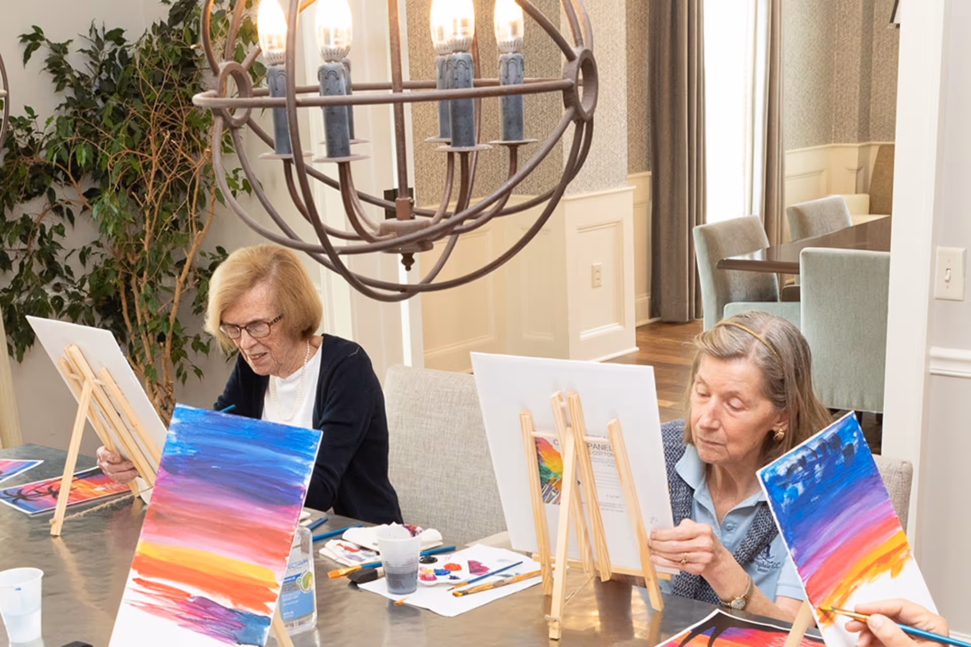 Two elderly women seated at a table in a well-lit room, engaged in painting colorful landscapes on canvases placed on wooden easels. The room has a large decorative hanging light fixture and a green leafy plant in the background.