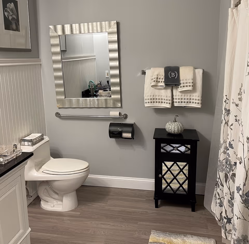 A clean and modern bathroom with a white toilet, a silver-framed mirror above a grab bar, a black cabinet with a decorative pumpkin on top, and a towel rack holding three neatly folded towels. The walls are painted light gray, and there is a floral shower curtain on the right side.